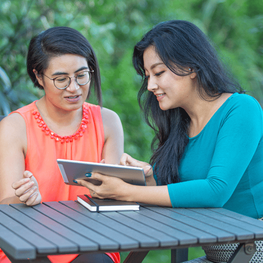 Two women sitting down at a park bench outside looking at a iPad 